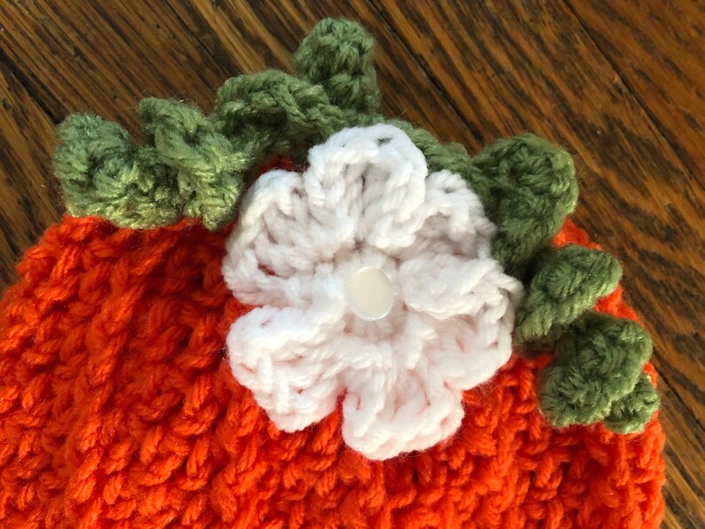 May include: Close-up of a handmade orange crochet pumpkin hat with a white flower and green leaves. The hat is made of a textured yarn and features a button in the center of the flower. Perfect for fall and Halloween.