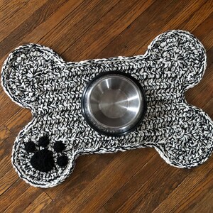 May include: Black and white crocheted dog bone shaped pet food mat with a paw print design and a stainless steel food bowl.