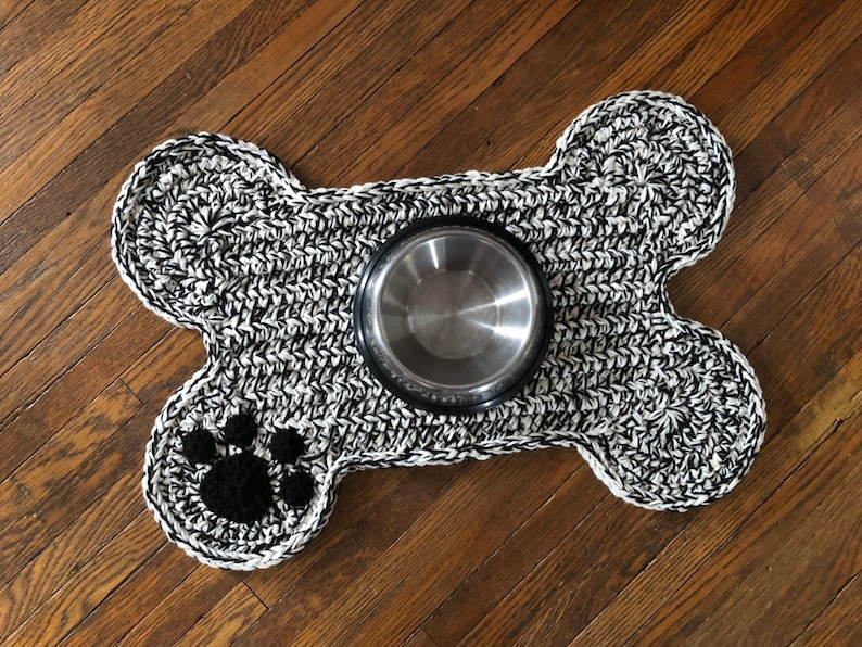 May include: Black and white crocheted dog bone shaped pet food mat with a paw print design and a stainless steel food bowl.