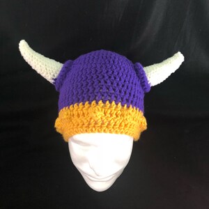 Pu&ograve; includere: Cappello all'uncinetto viola e oro con corna bianche. Il cappello &egrave; su una testa di manichino bianca.