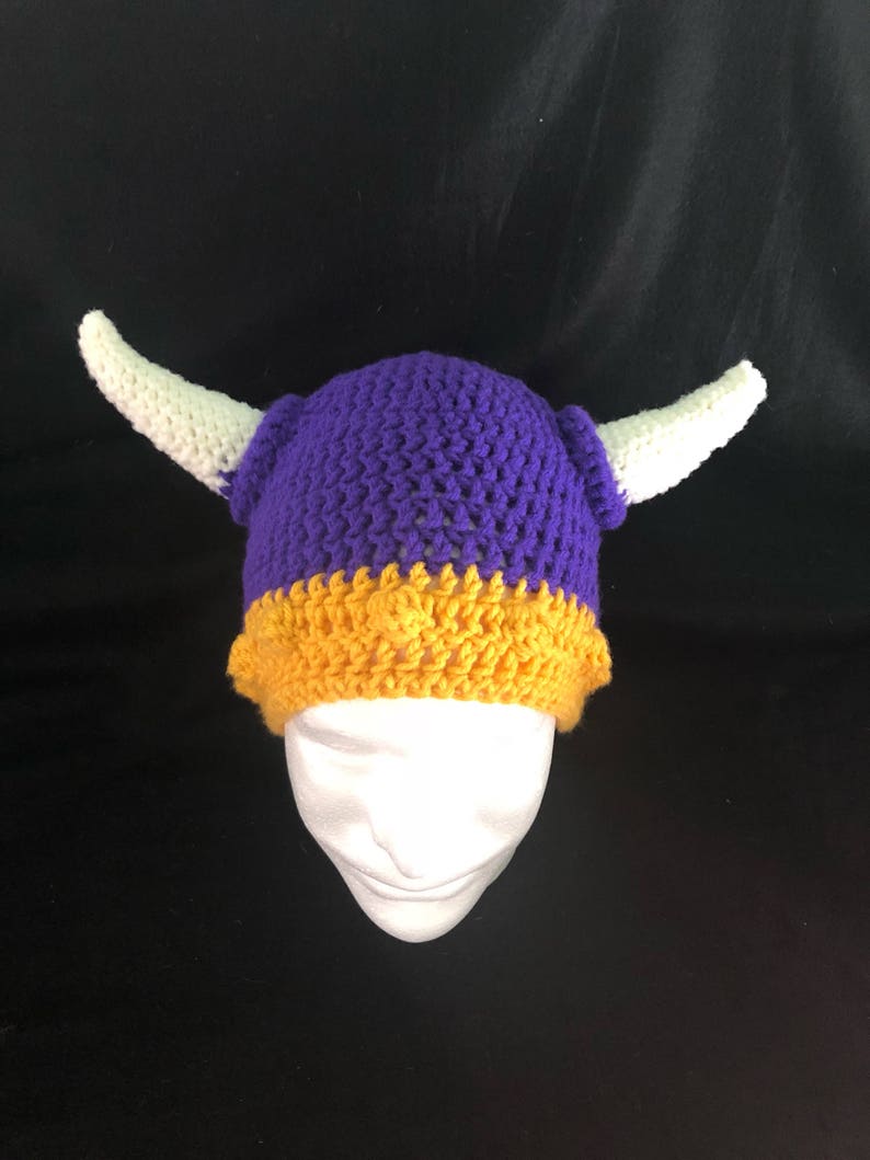 Pu&ograve; includere: Cappello all'uncinetto viola e oro con corna bianche. Il cappello &egrave; su una testa di manichino bianca.