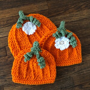 May include: Three orange crochet hats shaped like pumpkins, each with a green stem and white flower. The hats are arranged on a wooden surface, showcasing the handmade details and fall theme. The hats are perfect for a baby or toddler.