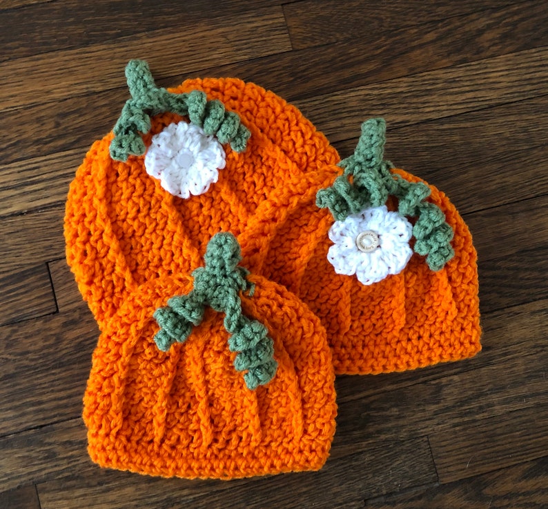 May include: Three orange crochet hats shaped like pumpkins, each with a green stem and white flower. The hats are arranged on a wooden surface, showcasing the handmade details and fall theme. The hats are perfect for a baby or toddler.