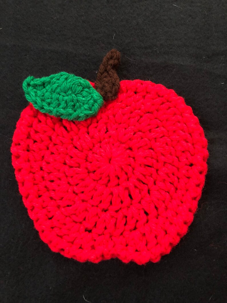 Puede incluir: Una manzana roja de ganchillo con una hoja verde y un tallo marr&oacute;n. La manzana est&aacute; hecha con punto bajo y tiene una superficie texturizada.