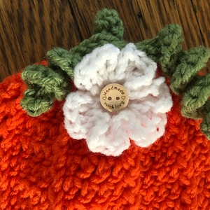 May include: Handmade orange crochet hat with a white flower and green leaves. A small wooden button in the center of the flower reads "handmade with love". The hat is a cozy, handcrafted item.