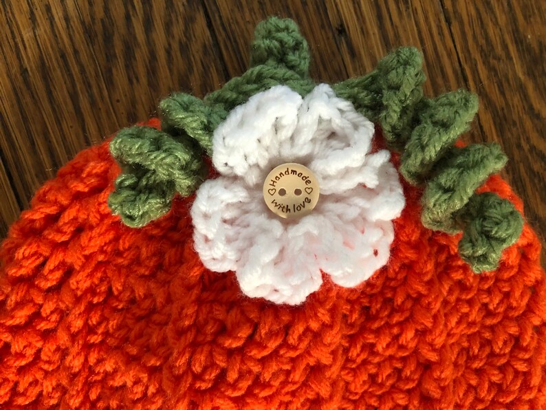 May include: Handmade orange crochet hat with a white flower and green leaves. A small wooden button in the center of the flower reads "handmade with love". The hat is a cozy, handcrafted item.
