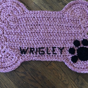 May include: A pink crocheted dog bone shaped mat with the name "WRIGLEY" embroidered in black thread and a black paw print.
