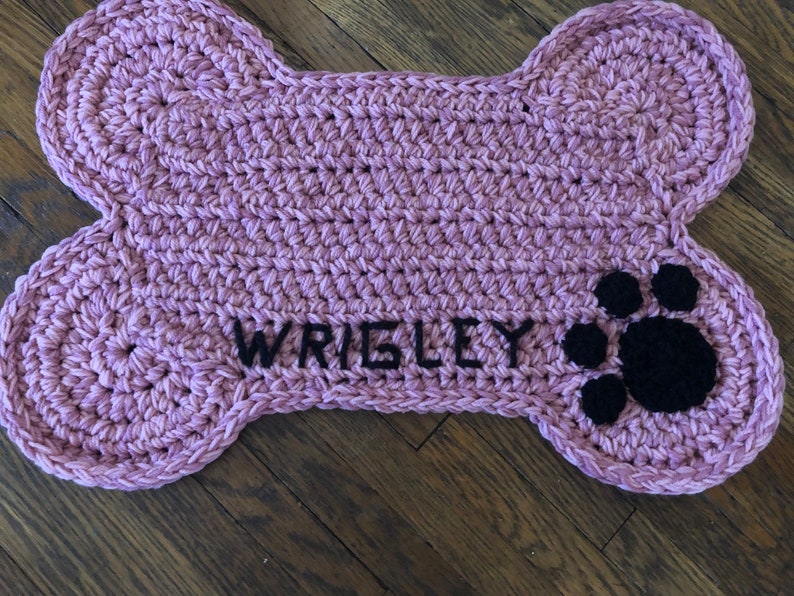 May include: A pink crocheted dog bone shaped mat with the name "WRIGLEY" embroidered in black thread and a black paw print.