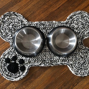 May include: Black and white crocheted dog bone shaped mat with two stainless steel bowls. The mat has a black paw print in the center.