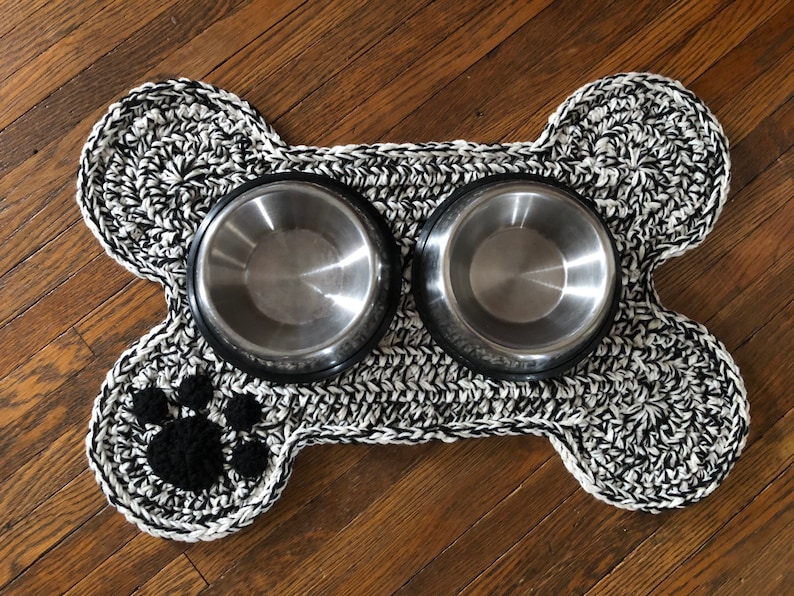 May include: Black and white crocheted dog bone shaped mat with two stainless steel bowls. The mat has a black paw print in the center.
