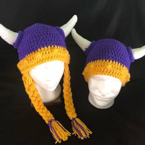 Pu&ograve; includere: Due cappelli all'uncinetto con corna bianche e trecce gialle. I cappelli sono viola e gialli.