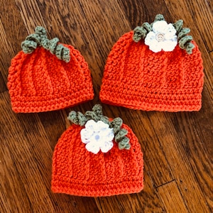 May include: Three orange crochet hats shaped like pumpkins, each with green leaves and a white flower. The hats are arranged on a wooden surface, showcasing the handmade details and autumnal theme.