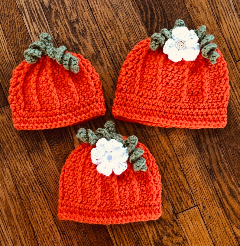 May include: Three orange crochet hats shaped like pumpkins, each with green leaves and a white flower. The hats are arranged on a wooden surface, showcasing the handmade details and autumnal theme.