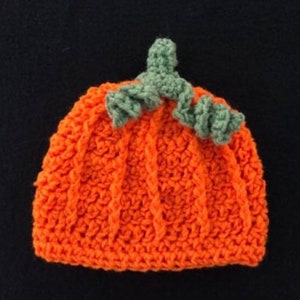 May include: An orange crochet hat shaped like a pumpkin, with vertical ribbing and a green stem and leaves. The hat is handmade and likely for a baby or child, perfect for Halloween or fall-themed photoshoots.