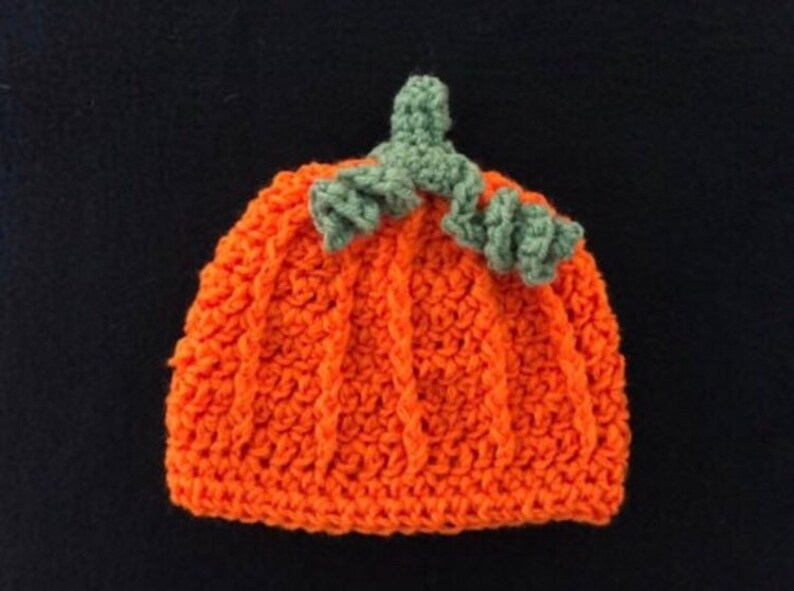 May include: An orange crochet hat shaped like a pumpkin, with vertical ribbing and a green stem and leaves. The hat is handmade and likely for a baby or child, perfect for Halloween or fall-themed photoshoots.