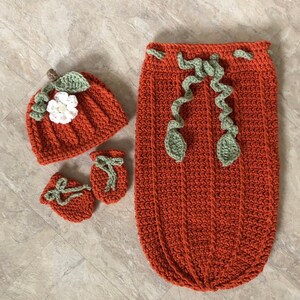 Op de afbeelding: Een gehaakte baby-outfitset in de vorm van een pompoen, in oranje, groen en wit. De set bevat een muts, slofjes en een slaapzak.