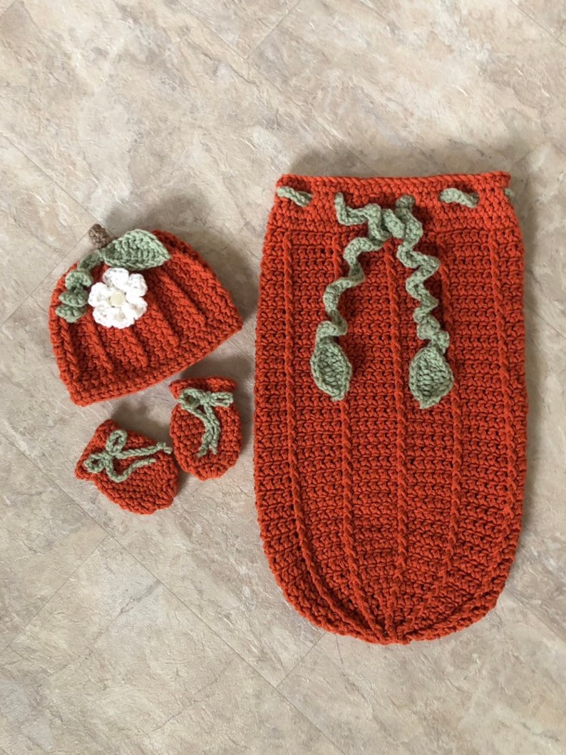 Op de afbeelding: Een gehaakte baby-outfitset in de vorm van een pompoen, in oranje, groen en wit. De set bevat een muts, slofjes en een slaapzak.