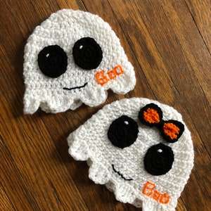 May include: Two white crocheted ghost hats with black eyes and a stitched-on smile. One hat has an orange bow and both hats have the word "Boo" stitched on them.