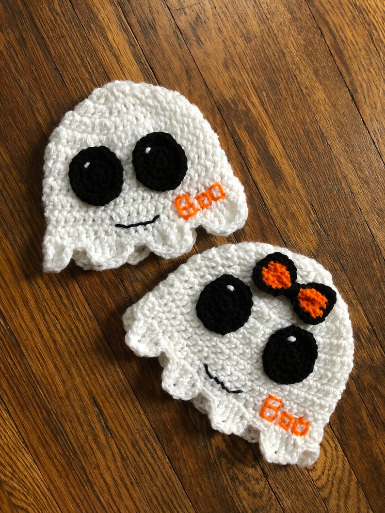 May include: Two white crocheted ghost hats with black eyes and a stitched-on smile. One hat has an orange bow and both hats have the word "Boo" stitched on them.