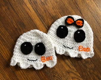 May include: Two white crocheted ghost hats with black eyes and a smiling mouth. One ghost has a small orange bow on top and both say "Boo" in orange yarn.