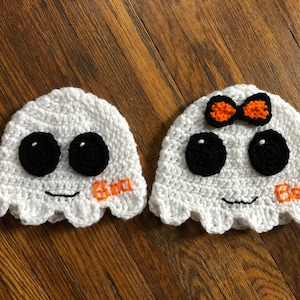 May include: Two white crocheted ghost hats with black eyes and a smiling mouth. One hat has a small orange and white bow on top. Both hats have the word "Boo" embroidered on the front.