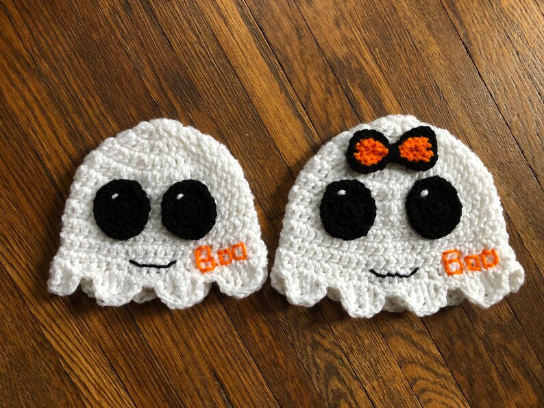 May include: Two white crocheted ghost hats with black eyes and a smiling mouth. One hat has a small orange and white bow on top. Both hats have the word "Boo" embroidered on the front.