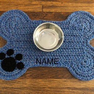 May include: Blue crocheted dog bone shaped mat with a black paw print and the word "NAME" embroidered on it. A silver metal bowl is on top of the mat.