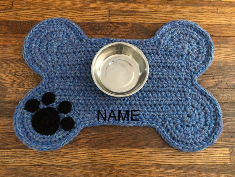 May include: Blue crocheted dog bone shaped mat with a black paw print and the word "NAME" embroidered on it. A silver metal bowl is on top of the mat.