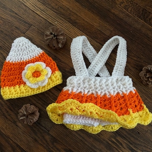 May include: A crocheted candy corn costume for a baby, featuring a hat and a dress with a white, orange, and yellow color scheme. The hat has a flower detail.