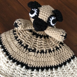 Op de afbeelding: Een gehaakte veiligheidsdeken in de vorm van een mops met een beige en zwart lichaam en een witte rand. De deken is gemaakt van zacht garen en is perfect om te knuffelen.