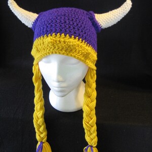 Pu&ograve; includere: Cappello all'uncinetto viola e giallo con corna bianche e trecce gialle. Il cappello ha uno schema di colori viola e giallo.