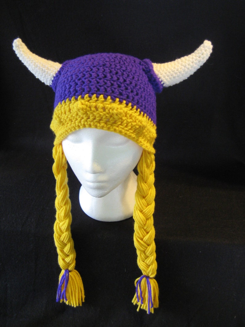 Pu&ograve; includere: Cappello all'uncinetto viola e giallo con corna bianche e trecce gialle. Il cappello ha uno schema di colori viola e giallo.