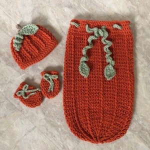 Op de afbeelding: Een gehaakte baby-outfitset met een pompoenthema, inclusief een muts, slofjes en een slaapzak. De set is gemaakt in tinten oranje en groen.