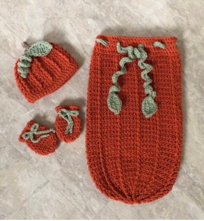 Op de afbeelding: Een gehaakte baby-outfitset met een pompoenthema, inclusief een muts, slofjes en een slaapzak. De set is gemaakt in tinten oranje en groen.