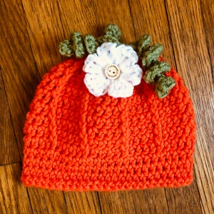 May include: An orange crochet hat, resembling a pumpkin, with a white flower and green leaves on top. The flower has a wooden button center. The hat is handmade and suitable for babies or toddlers.