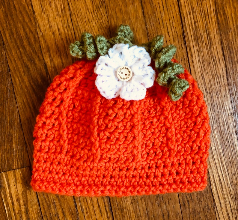 May include: An orange crochet hat, resembling a pumpkin, with a white flower and green leaves on top. The flower has a wooden button center. The hat is handmade and suitable for babies or toddlers.