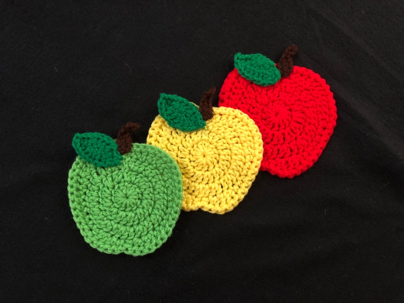 Puede incluir: Tres posavasos de ganchillo con forma de manzana en rojo, amarillo y verde. Cada posavasos tiene una hoja verde y un tallo marr&oacute;n.