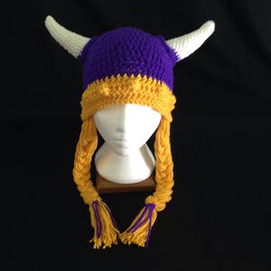 Pu&ograve; includere: Cappello vichingo all'uncinetto viola e oro con corna bianche e lunghe trecce gialle.