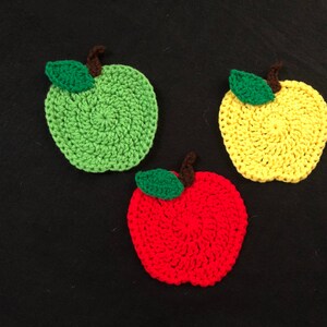 Puede incluir: Tres posavasos de ganchillo con forma de manzana. Uno es verde, uno es amarillo y uno es rojo.