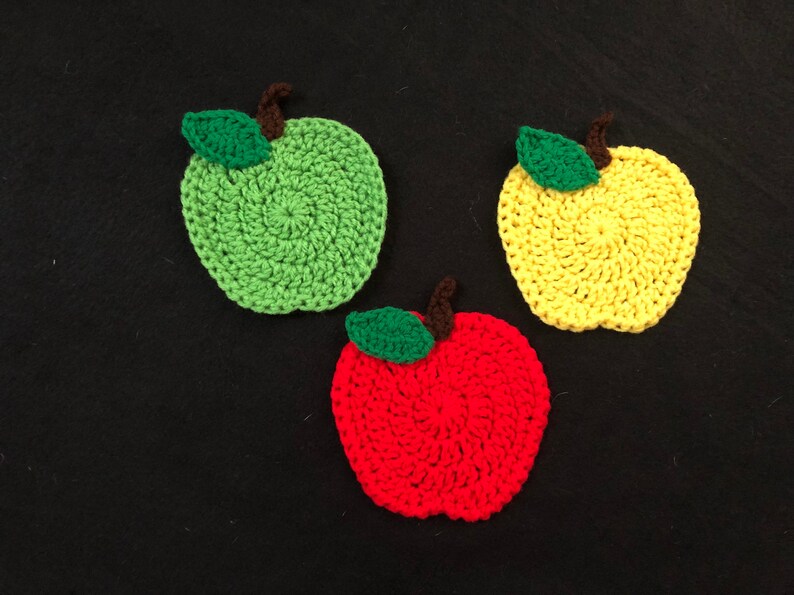 Puede incluir: Tres posavasos de ganchillo con forma de manzana. Uno es verde, uno es amarillo y uno es rojo.
