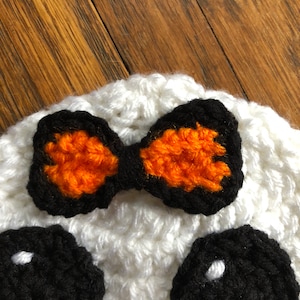 May include: White crocheted hat with black eyes and an orange and black bow. The hat is a fun and festive accessory for Halloween.
