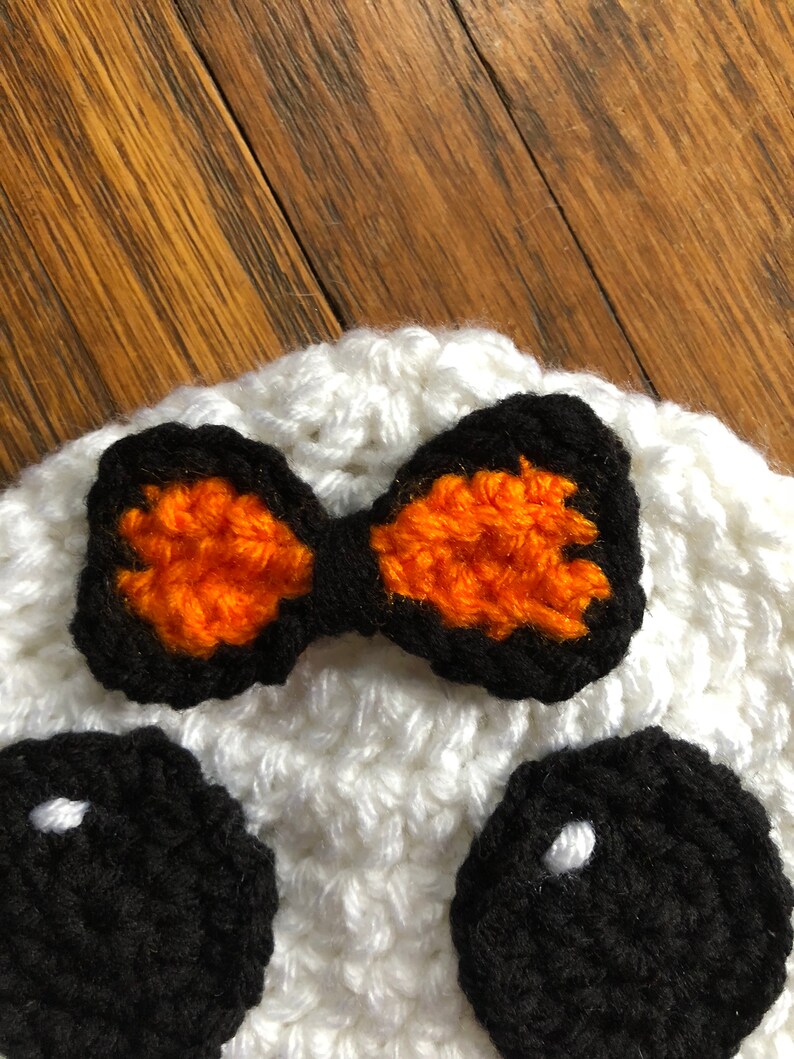 May include: White crocheted hat with black eyes and an orange and black bow. The hat is a fun and festive accessory for Halloween.