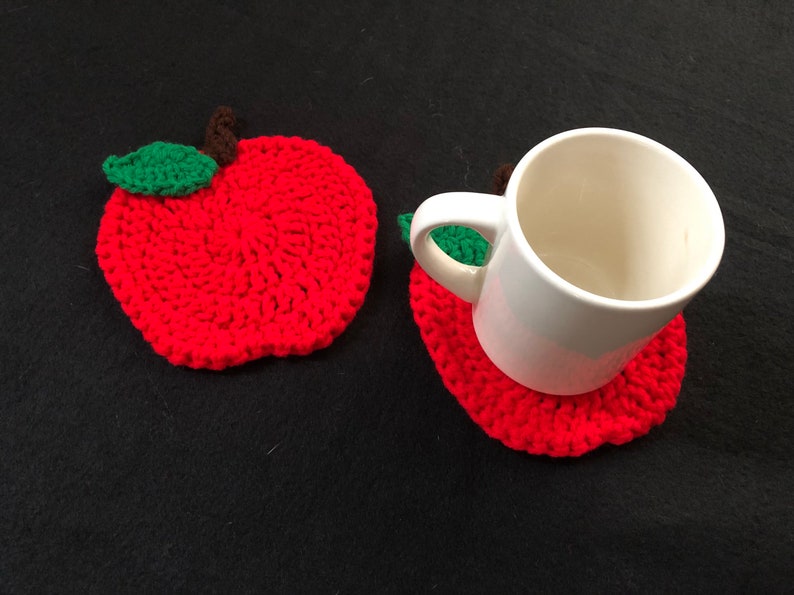 Puede incluir: Dos posavasos de ganchillo en forma de manzana roja con hojas verdes. Un posavasos tiene una taza blanca encima.