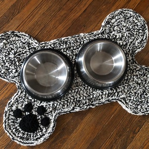 May include: A black and white crocheted dog bone shaped mat with two stainless steel bowls. The mat has a black paw print in the center.