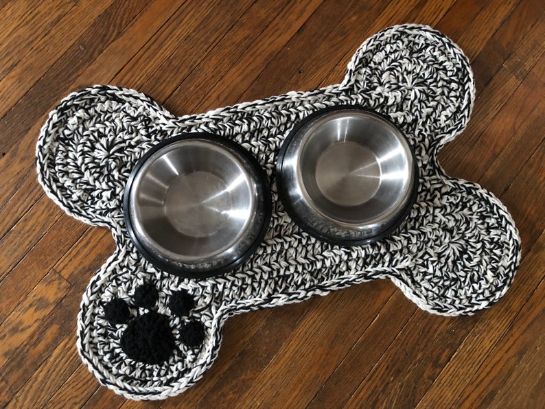 May include: A black and white crocheted dog bone shaped mat with two stainless steel bowls. The mat has a black paw print in the center.