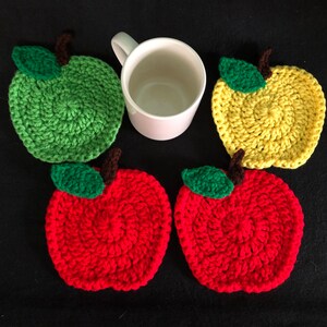 Puede incluir: Cuatro posavasos de ganchillo con forma de manzana en rojo, verde y amarillo. Los posavasos est&aacute;n dispuestos en c&iacute;rculo alrededor de una taza blanca.