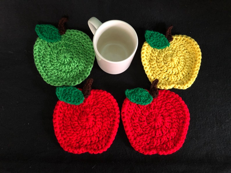 Puede incluir: Cuatro posavasos de ganchillo con forma de manzana en rojo, verde y amarillo. Los posavasos est&aacute;n dispuestos en c&iacute;rculo alrededor de una taza blanca.