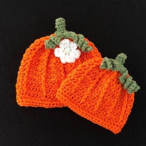 May include: Two orange crochet pumpkin hats with green stems and white flower accents. The hats are handmade and have a ribbed texture, perfect for fall or Halloween. The hats are likely for babies or small children.