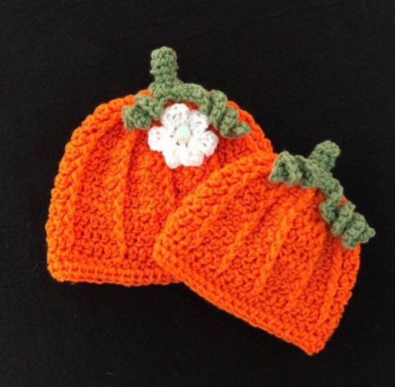 May include: Two orange crochet pumpkin hats with green stems and white flower accents. The hats are handmade and have a ribbed texture, perfect for fall or Halloween. The hats are likely for babies or small children.