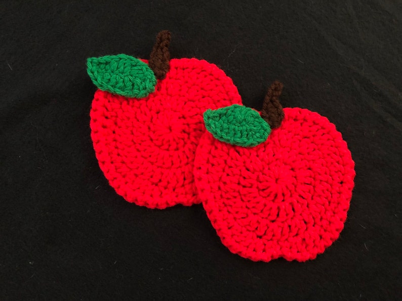 Puede incluir: Dos posavasos de ganchillo de color rojo en forma de manzana con hojas de ganchillo verdes y tallos marrones.
